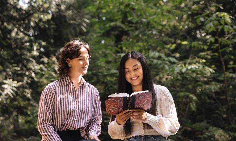 Two female students standing in front of greenery and trees reading the Bible and smiling.