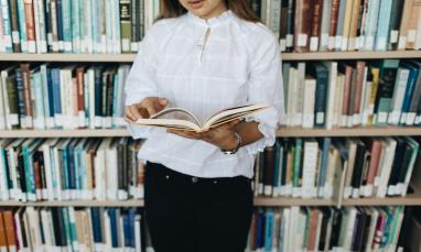 Student holding an open book in the library in front of a shelf of books.
