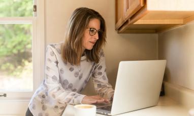 Woman in kitchen working on laptop while standing at a cupboard
