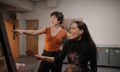 A student holding a paintbrush, with an instructor pointing at her work and smiling