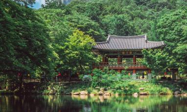 A traditional Korean pavilion with a dark-tiled roof and wooden pillars stands by a tranquil pond, surrounded by lush green trees. The water reflects the pavilion and the surrounding foliage, creating a mirror-like effect. In the background, a towering mountain covered in dense vegetation rises under a partly cloudy sky. Small colorful lanterns hang along a wooden fence, adding a festive touch to the serene natural setting.