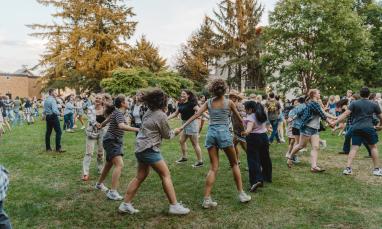 A group of people holding hands and dancing in a circle on a grassy field during an outdoor event.