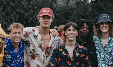A group of seven people in tropical-themed outfits, including floral shirts and grass skirts, pose outdoors; one is dressed in a banana costume.