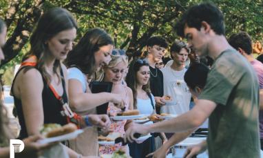 Young men and women standing in line at an outdoor buffet, serving themselves food in a park-like setting with trees in the background.