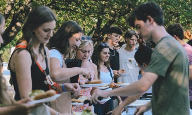 Young men and women standing in line at an outdoor buffet, serving themselves food in a park-like setting with trees in the background.