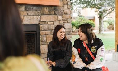 Two young women sit in front of a stone fireplace, smiling at each other. One holds a boba drink with a purple straw. A third person is partially visible in the foreground.