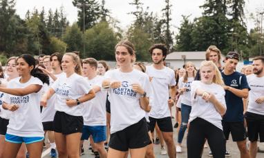 A group of young adults in white "SPARTANS TOGETHER" t-shirts participate in a synchronized outdoor activity, with trees and a building in the background.