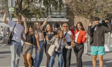 Group of nine young men and women posing casually in a parking lot with trees and a building in the background.