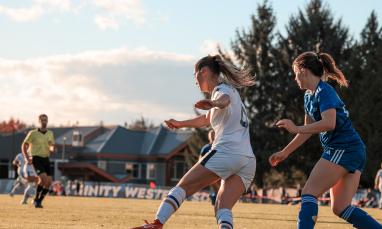 Two soccer players, one in white and one in blue, compete for the ball on an outdoor field, with a referee and teammates in the background. A "TRINITY WEST" banner is visible on the sideline.