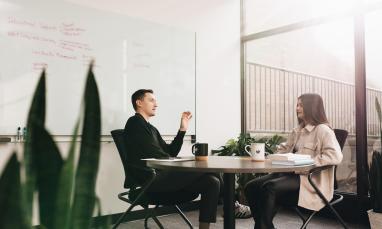 Two people sit at a round table in an office, talking, with a whiteboard behind them.