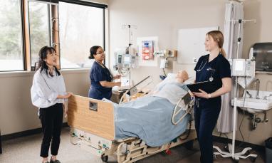 Three people stand around a patient in a hospital bed, surrounded by medical equipment and natural light from nearby windows.
