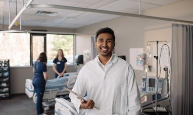 A person in a white lab coat holds a clipboard in a hospital room, while two others stand near a bed surrounded by medical equipment.