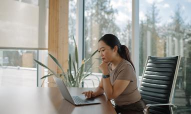 A young woman sits at a desk in front of a laptop, one hand on the keyboard and the other resting on their chin.