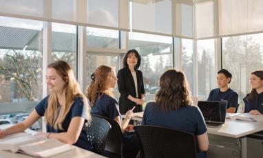 A group of students sit around a classroom table with notebooks and laptops while an instructor stands at the head of the table.