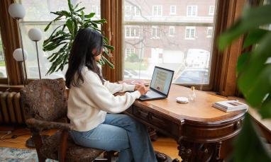  Person in a white sweater working on a laptop at a wooden desk near large windows with a city view.