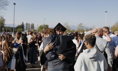A person in a graduation cap and gown hugs someone during an outdoor ceremony, surrounded by people with flowers and gifts, with trees and mountains in the background.