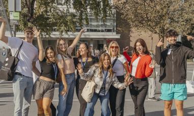 Group of nine young men and women posing casually in a parking lot with trees and a building in the background.