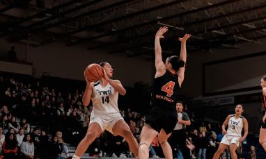 TWU basketball player in white jersey number 14 holds the ball while being closely defended by a player in black jersey number 4.
