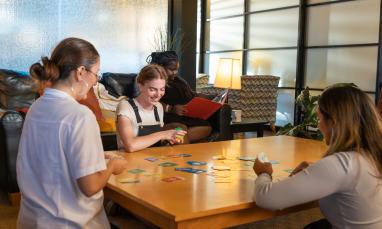 Three women play a card game at a table, one reads on a couch nearby.