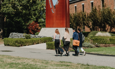 Three people walking on campus path near a brick building and red sculpture.
