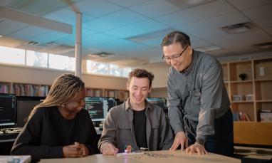 Three people smiling and collaborating in a library with a circuit board on the table.
