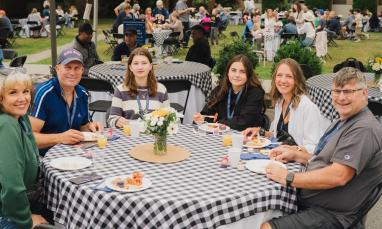 Group of people sitting at an outdoor table with checkered tablecloths, smiling.