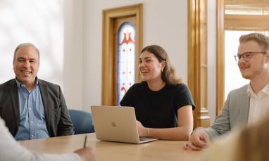 Three people smiling around a table, one with a laptop, in a well-lit room.