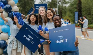 Four people smiling, holding blue signs saying "YOU MADE IT" with balloons nearby.