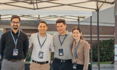Four people smiling under a canopy with lanyards, next to a sign.
