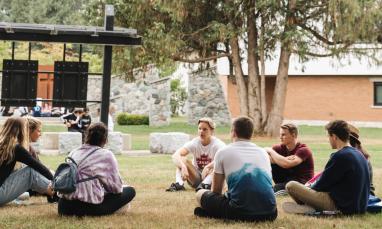 Students sitting in a circle on grass, school building in background.