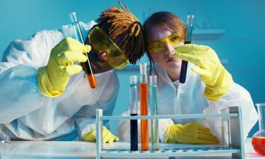 Two scientists in lab coats examine test tubes with serious expressions.