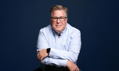Smiling man with glasses in a blue shirt against a dark blue background.