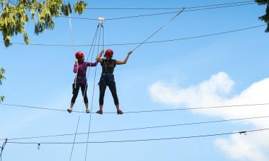 Two people on a rope course in a forest, wearing helmets, under a blue sky.