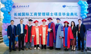 Graduates and officials in robes pose at a ceremony, with blue balloons and a banner backdrop.