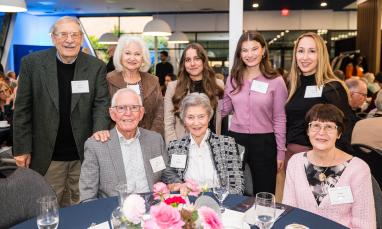 Group of people of various ages smiling at an event, gathered around a table with flowers.