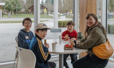 Woman and three young boys sitting at a table with paper cup drinks, playing cards, facing the camera with a smile.