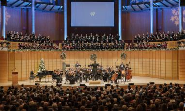 A full orchestra performing on stage and a conductor leading them during the Christmas concert. Behind the orchestra, a large choir stands on an elevated platform, and the stage is decorated with wreaths, garlands, and a Christmas tree, creating a festive atmosphere.