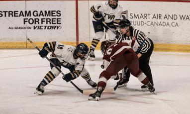 Two hockey players face off on the ice, one in a white TWU jersey and the other in a maroon uniform, with a referee dropping the puck and another player in white positioned behind them. Advertising boards are visible in the background.