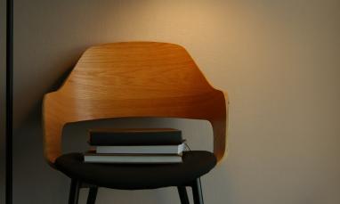A wooden chair with a black cushioned seat sits against a neutral wall, illuminated by soft lighting. A small stack of books rests neatly on the seat.