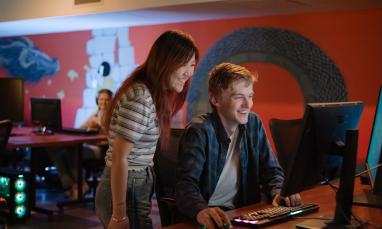 Two people work together at a computer station in a dimly lit room with a colorful mural on the wall. 