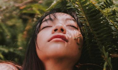 A woman with long dark hair is positioned outdoors among dense, green fern leaves. 