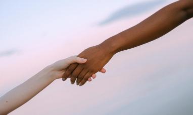 Two hands reaching toward each other and holding against a soft, pastel sky background.