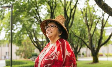 Patti Victor Switametelót in her cultural regalia smiling and looking past the camera in front of greenery