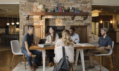 TEDS students gather around a table in the Graduate Collegium, participating in a discussion with laptops, books, and notebooks in a lounge-style learning space featuring a large stone fireplace.