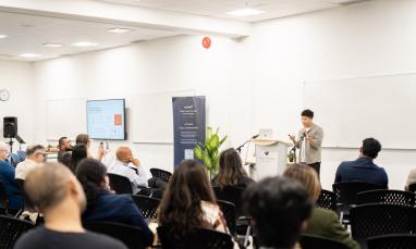 Audience members seated in a classroom-style event space at TWU Richmond campus listen as a speaker presents from a podium. A presentation screen, TWU-branded banner, and whiteboards are visible at the front of the room.