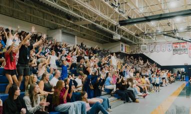 A crowd of adults and children cheer with raised hands from gymnasium bleachers during TWU Homecoming.