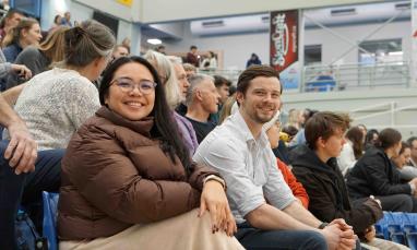 Two attendees smiling at the camera while seated among spectators in gymnasium.