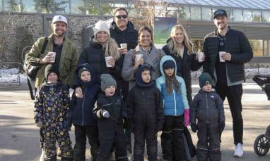 A group of adults and children dressed for cool weather gather outdoors in front of a campus building while holding warm drinks.