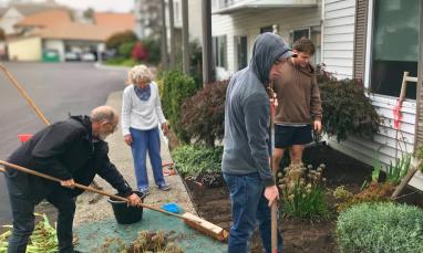 group of people working together outdoors on a community service project, using rakes and shovels around a garden area.