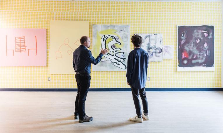 A professor and a student talking while looking at a wall displaying five art pieces.